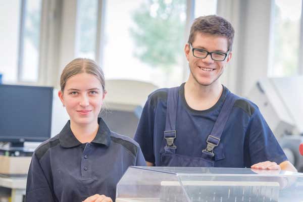 Eine junge Frau und ein junger Mann stehen freundlich lächelnd in der Produktionshalle einer Druckerei